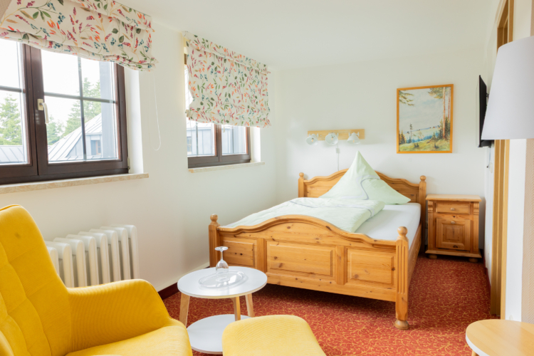 Ein Zimmer in dem ein Bett aus Holz steht. Der Fußboden ist rot und ein gelber Sessel mit kleinem Beistelltisch ist zu sehen.